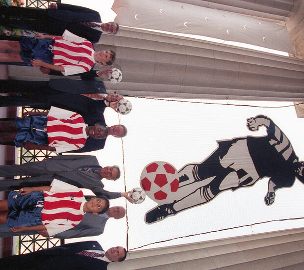 This eye-level group portrait captures President Bill Clinton (center) during the 1994 FIFA World Cup opening ceremony at Soldier Field in Chicago. Standing alongside the President are several high-ranking officials and young soccer fans. Three young soccer players stand in the foreground, wearing the official 1994 USA home jersey featuring iconic red-and-white stripes. The group is framed by the stadium's historic Doric columns, with a massive banner of a stylized soccer player suspended behind them.