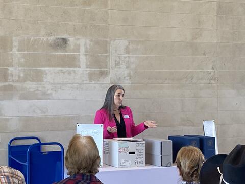  A woman with long graying hair, wearing a vibrant pink blazer over a dark top. She is gesturing with her hands while addressing the crowd. In front of her are several archive-style storage boxes. One prominent white box has handwritten labels, including numbers like "5285" and "8102". Two dark blue boxes or cases sit to the right.