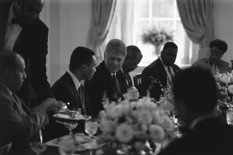 Black and White Photograph. President Clinton sits next to Rep. Kweisi Mfume. The two men are speaking to each other. Other members of the Congressional Black Caucus can be seen sitting around Clinton and Mfume.
