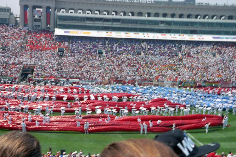 An eye-level shot captures a massive crowd at a stadium, where a sprawling red and blue fabric display covers much of the field. Dozens of people in white uniforms stand amidst the fabric, holding white poles that appear to support the display. In the background, the stadium stands are packed with spectators, and a large multi-story building with flags on top rises behind the seating area. The overall scene is bright.