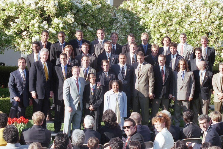 An eye-level, medium-wide shot captures Bill Clinton posing for a group photograph with the 1996 D.C. United team and staff. Clinton, wearing a navy suit and gold tie, stands on the left side of the second row.The large group, primarily composed of men in various suits and ties, is arranged in multiple rows on an outdoor stage. In the front row, a young boy and girl stand centrally. The group is set against a backdrop of lush green shrubbery and white flowering trees. 