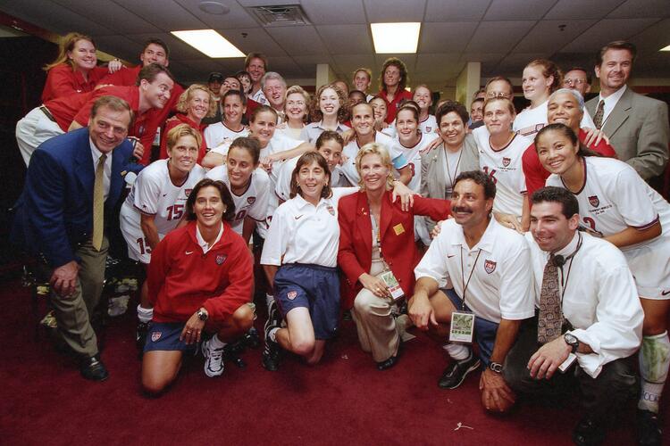 The crowded group poses closely together on a red carpet. Former President Bill Clinton is visible near the back center, smiling alongside Hillary Clinton and Chelsea Clinton. Surrounding them are numerous USWNT soccer players in white jerseys, coaches, and staff members smiling at the camera. Several men in suits and athletic staff in white polos frame the edges of the group..