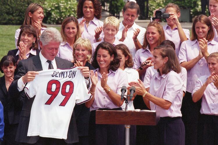 An outdoor White House ceremony features Bill Clinton holding up a white soccer jersey with the name "CLINTON" and the number "99" printed on the back. He stands on the left in a dark pinstripe suit. Two female soccer players stand next to him behind a wooden podium, smiling and clapping. A large group of team members, dressed in matching light purple button-down shirts, are gathered closely behind them, clapping and cheering in bright daylight.