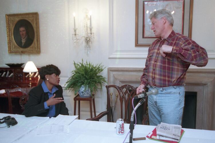 President Clinton Stands in blue jeans. Minyon Moore sits behind a table covered in white table cloth.
