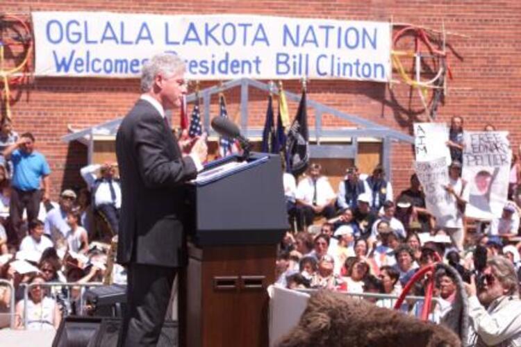 President Clinton stands behind the Presidential Podium. A sign in the back reads Oglala Lakota Nation Welcomes President Bill Clinton.