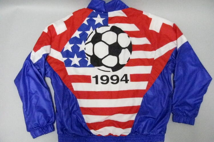 The back view of a vintage tracksuit jacket is laid flat, featuring a bold red, white, and blue graphic design celebrating the 1994 World Cup. The center panel displays a large black and white soccer ball set against a background mimicking the American flag, with white stars on a blue field to the left and horizontal red and white stripes across the rest of the panel. The year "1994" is printed in large black numbers directly beneath the soccer ball. The jacket has solid blue sleeves.