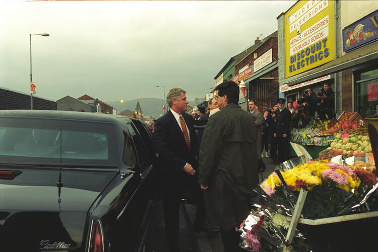 President Clinton exits his motorcade. He stands by the a black Presidential Limo. A crowd is gathered.