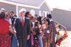 Title President Clinton with Visitors and Residents of Pine Ridge Reservation President Clinton stands in a suit. President Clinton shakes a resident's hand. The croup of residents stand in front of a home that is painted grey. A young person can be seen in the background examining a ceremonial headdress.