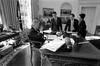 Gene Sperling, Rahm Emanuel, and Alexis Herman stand in the Oval Office briefing President Clinton as he sits behind his desk, with his hands touching in front of his face. 