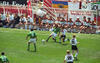 An action shot from a soccer match shows a player in a green jersey (number 16) controlling the ball on a sunny grass field. He is pursued by German players in white jerseys and black shorts, including (number 16) and (number 20). In the background, the German substitute bench sits in a transparent dugout, with various banners and a German flag visible behind them.