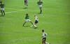 In this action shot from an international soccer match, a German player in a white jersey and black shorts (number 16), battles for the ball on a sunny grass field. He is closely marked by an opposing player in a green jersey, likely from Bolivia, who also wears the number 16. In the foreground, another German player (number 14), runs toward the play, while other players in green jerseys are visible in the background.