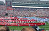 An eye-level shot captures a massive crowd at a stadium, where a sprawling red and blue fabric display covers much of the field. Dozens of people in white uniforms stand amidst the fabric, holding white poles that appear to support the display. In the background, the stadium stands are packed with spectators, and a large multi-story building with flags on top rises behind the seating area. The overall scene is bright.