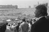 This black-and-white photograph shows President Bill Clinton in profile, overlooking the 1994 FIFA World Cup opening ceremony at Soldier Field. In the foreground, Clinton stands in a dark suit, his gaze directed toward the field where performers are arranged in large formations. The stadium's stands are overflowing with spectators, and in the distance, a large electronic scoreboard displays the text "WorldCupUSA94." The Chicago skyline, including the Willis Tower (then the Sears Tower), is visible.