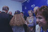 In the center, a person (Chelsea Clinton) with long, curly blonde hair wearing a purple baseball cap stands with their back to the camera. To their right, an older woman with short blonde hair, wearing glasses and a tan blouse with a prominent event credential, smiles toward someone off-camera. Several men in suits are partially visible in the background and foreground, including Bill Clinton, whose back is to the camera on the left. The lighting is slightly dim, typical of an indoor event space.