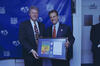 This eye-level photograph shows President Bill Clinton and Alan Rothenberg, then-President of U.S. Soccer, smiling as they pose together at the 1994 FIFA World Cup. Both men are dressed in dark suits; Clinton wears a colorful patterned tie, while Rothenberg wears a solid red tie. Together, they hold a large framed commemorative display featuring the official tournament poster, several silver coins, and an official document. They stand in front of a blue backdrop emblazoned with the WorldCupUSA94 and FIFA.