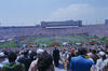 1994 FIFA World Cup opening ceremony at Soldier Field in Chicago. The stadium is filled to capacity with a dense crowd of spectators under a bright, partly cloudy sky. On the green field below, hundreds of performers in white uniforms are arranged in a large, circular formation, surrounded by vibrant displays of colorful fabrics and various international flags. In the foreground, the backs of spectators in the stands are visible, including a man in a dark suit standing prominently in the center aisle. 