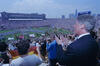 President Bill Clinton (on the right) looking out and applauding from the stands during the 1994 FIFA World Cup opening ceremony at Soldier Field. In the foreground, the backs and sides of various spectators are visible, including one person holding a small flag. The stadium is packed with a massive crowd. On the green field, hundreds of performers in white uniforms are organized into large formations, accompanied by colorful banners and flags, including the flag of South Korea. 