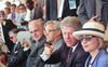 President Bill Clinton is shown speaking and gesturing toward the field while wearing a dark suit and a colorful patterned tie.Hillary Rodham Clinton: The First Lady sits next to him, wearing a blue-and-white checkered blazer and a wide-brimmed cream hat.Helmut Kohl: The former German Chancellor is seated to the President's right (on the left side of the frame), wearing large, thin-framed glasses.