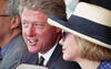 On the left, President Clinton is shown in a three-quarter view, smiling and looking toward the field. He wears a dark suit and a colorful patterned tie. To his right, Hillary Clinton is seen in profile, wearing a blue checkered blazer and a wide-brimmed cream hat with a black band. A third person, partially visible on the far left, wears glasses and a gray suit. The lighting is bright and natural, typical of an outdoor afternoon event.