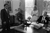 Black and white photograph. President Clinton meets with Director of the Office of Scheduling and Advance, Stephanie Streett, and President's Aide, Stephen Goodin in the White House Oval Office. Goodin and Streett are standing, President Clinton sits behind his desk. All are smiling.
