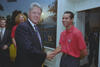 A medium shot shows U.S. President Bill Clinton shaking hands with soccer player Roy Wegerle inside the White House. On the left, Clinton wears a dark suit, white shirt, and blue patterned tie while smiling at the camera. On the right, Pérez wears a red USA soccer polo shirt and dark pants, smiling back at Clinton. Two men stand in the background on the left, and a scenic landscape painting hangs on the wall behind them.