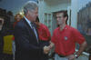 A close-up shot capturing President Bill Clinton shaking hands with Mike Burns. Burns wears a red polo shirt adorned with a U.S. soccer logo. President Clinton, on the left, is seen smiling warmly as he extends his hand. Burns is on the right, also smiling, returns the handshake. In the background, other individuals in matching red shirts are visible, along with a decorated doorway and a mural depicting a natural landscape.