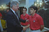 A medium shot capturing President Bill Clinton shaking hands with Claudio Reyna who wears a red polo shirt featuring a U.S. soccer logo. President Clinton is on the left, smiling, while another man in an identical red polo shirt stands directly behind them, looking on. The setting features a decorated room with scenic landscape wallpaper and an ornate wall sconce lighting fixture visible on the right.