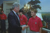 A medium shot capturing President Bill Clinton on the left, smiling as he shakes hands with a blonde man wearing a red U.S. soccer team polo shirt. In the background, two other team members in identical red shirts look on, with one standing partially behind the President. The room is decorated with a scenic nature mural on the wall and an elegant crystal wall sconce light fixture.