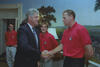 A medium shot capturing President Bill Clinton on the left, smiling as he shakes hands with a tall, short-haired man wearing a red U.S. soccer team polo shirt. In the background, two other team members in identical red shirts look on, with one standing directly between the President and the player. The room features a scenic nature mural on the wall and a white table lamp visible behind the men.