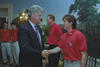 A medium shot capturing President Bill Clinton on the left, smiling as he shakes hands with a man with brown hair wearing a red U.S. soccer team polo shirt. In the background, three other team members in identical red shirts look on, with one standing directly between the President and the player. The room features a scenic nature mural on the wall, an ornate light fixture, and wooden chairs.