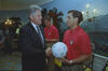 A medium shot capturing President Bill Clinton on the left, smiling as he shakes hands with a man with short brown hair wearing a red U.S. soccer team polo shirt. The man holds a white soccer ball covered in signatures. In the background, another team member looks down, and a man in a dark suit stands on the far left. The room features a scenic nature mural on the wall, an ornate wall sconce on the right, and a grandfather clock.
