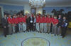 A wide group shot capturing President Bill Clinton and First Lady Hillary Clinton standing in the center with members of the U.S. Men's National Soccer Team. President Clinton holds a white soccer ball while surrounded by players and staff wearing red team polo shirts and khaki pants. A young Chelsea Clinton stands to the left of her parents. They are all gathered in a semi-circle inside a grand, oval-shaped room featuring a large crystal chandelier overhead, a patterned circular rug on the floor.