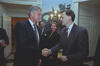 A photograph captures President Bill Clinton shaking hands with a man in a suit inside a White House room. Clinton stands on the left, looking toward the man on the right, who is smiling slightly. In the background between them, a man with a mustache and glasses and a woman with blonde hair look on and smile. They are standing in front of a wall with scenic wallpaper depicting trees and a stone bridge, lit by a wall sconce.