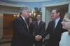 A photograph captures President Bill Clinton shaking hands with a man wearing glasses and a dark suit inside a White House room. Clinton stands on the left, smiling as he grips the man's hand. To the right, a young woman wearing glasses is seen in partial profile, looking toward the interaction. In the background, a woman with blonde hair and the man from the previous photograph stand smiling. They are positioned in front of a wall with scenic wallpaper and a decorative wall sconce.