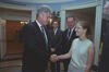 A photograph captures President Bill Clinton smiling as he shakes hands with a young woman wearing glasses and a light-colored sleeveless top. Clinton stands on the left in a dark suit and patterned tie. The young woman stands on the right, looking back at him with a smile. In the background between them, two men in suits look on—the man on the right wears glasses and smiles, while the man in the center watches the handshake. They are all gathered in a White House room featuring scenic wallpaper.