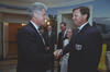 A medium shot capturing President Bill Clinton on the left, smiling as he shakes hands with a man wearing a dark blazer with a U.S. Soccer patch on the pocket. In the background between them, Chelsea Clinton stands smiling, wearing glasses and a sleeveless top, alongside two men in dark suits and patterned ties. The room features a scenic nature mural on the wall, an open doorway revealing a lit chandelier in an adjacent hallway, and a yellow upholstered chair visible on the far right.