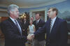 A photograph captures President Bill Clinton smiling as he shakes hands with a man wearing glasses and a striped tie. Clinton stands on the left in a dark suit with a patterned blue tie. The man stands on the right, looking toward Clinton while shaking his hand. In the background, several individuals look on, including a man in a dark blazer with a red logo badge, a young woman wearing glasses and a sleeveless top, and two other men in suits. They are gathered in a White House room with light blue walls.