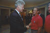 A photograph captures President Bill Clinton smiling as he shakes hands with a man wearing a red polo shirt. Clinton stands on the left in a dark suit with a patterned tie. The man stands on the right, looking back at Clinton with a slight smile; his red shirt features a USA crest on the left chest. In the background, several people look on from across a large room with blue walls, patterned carpeting, and scenic wallpaper.