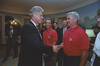 A photograph captures President Bill Clinton smiling as he shakes hands with an older man with grey hair wearing a red polo shirt. Clinton stands on the left in a dark suit. The man stands on the right, looking toward Clinton; his red polo features a USA crest on the left chest. In the background between them, the man from the previous photograph stands watching in an identical red polo shirt. Other onlookers in suits stand behind them in the White House room, which features scenic wallpaper.