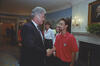 A photograph of President Bill Clinton shaking hands with a young man in the White House. Clinton, seen in profile wearing a dark suit and patterned tie, smiles as he grasps the hand of the man, who wears a red polo shirt featuring a U.S. Soccer logo. In the background, several onlookers smile, and a portrait of George Washington hangs above a nearby fireplace.