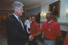 8A medium shot capturing President Bill Clinton on the left, smiling as he shakes hands with a Black soccer player wearing a red U.S. soccer team polo shirt. Another team member with a goatee stands directly behind the player, looking toward the camera. In the background, two women in white collared shirts stand near a scenic nature mural. The room features a white fireplace mantel, an oil portrait of George Washington, and ornate wall sconces.
