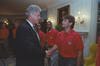 A photograph of President Bill Clinton shaking hands with a man wearing a red U.S. Soccer polo shirt inside the White House. Clinton, in profile on the left, wears a dark suit and patterned tie. The man looks directly at him as they shake hands. In the background, other members of the U.S. soccer team look on, and a framed portrait of George Washington hangs on the wall.