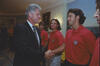 President Bill Clinton shaking hands with US Men's National Soccer Team player inside the White House. Clinton, shown in profile on the left wearing a dark suit and a blue patterned tie, smiles warmly. He standing on the right wearing a red U.S. Soccer polo shirt, looks toward the President. In the background between them, teammates watch the interaction, with a framed portrait of George Washington visible on the wall behind them.