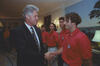 President Bill Clinton shaking hands with US Men's National Soccer Team player inside the White House. Clinton, shown in profile on the left wearing a dark suit and a blue patterned tie, smiles warmly. The player standing on the right wearing a red U.S. Soccer polo shirt, looks toward the President. In the background between them, teammates.