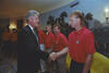 A medium shot captures former U.S. President Bill Clinton smiling and shaking hands with a man with a mullet hairstyle. The man and three others standing in line behind him wear matching red Nike polo shirts with a "USA" crest and khaki pants. The greeting takes place inside a room featuring a large, scenic wall mural of trees and a landscape, with several other individuals looking on in the background.