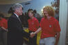 A medium shot capturing President Bill Clinton on the left, smiling as he shakes hands with a soccer player featuring long, curly blonde hair and a goatee. The player wears a red U.S. soccer team polo shirt. In the background between them, another teammate with lighter hair looks down and smiles. The room features a scenic nature mural on the wall, an ornate light fixture, and a bright yellow upholstered sofa behind the men.