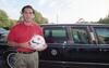 A medium shot capturing a soccer player standing outdoors on the left, holding a white soccer ball covered in signatures. He wears a red U.S. soccer team polo shirt and khaki pants. Behind him sits a black presidential limousine featuring the seal of the President of the United States on the door. In the distant background, the Washington Monument rises against a pale sky flanked by green trees.
