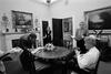 Black and white photograph. President Clinton meets with Stephanie Streett, Stephen Goodin and Nancy Hernreich in the Oval Office Dining Room. Heinreich and Streett stand while the President sits drinking a soft drink. Goodin stands leaning over the dining table.  