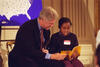 America Reads Event President Clinton sits with a child. They both hold a yellow book together. The child is wearing a name tag. There is a map of the united states in the background