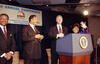 President Clinton laughs with Rev. Jesse Jackson, Sec. Alexis Herman. They all stand behind a podium.